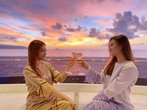 Two women sitting on a balcony of Party Boat Kota Kinabalu at sunset, clinking glasses.